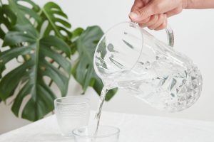 Woman's hand pouring fresh and pure water from a pitcher into a glass at the table. Drink water, healthy lifestyle. Concept of thirst and hydration.